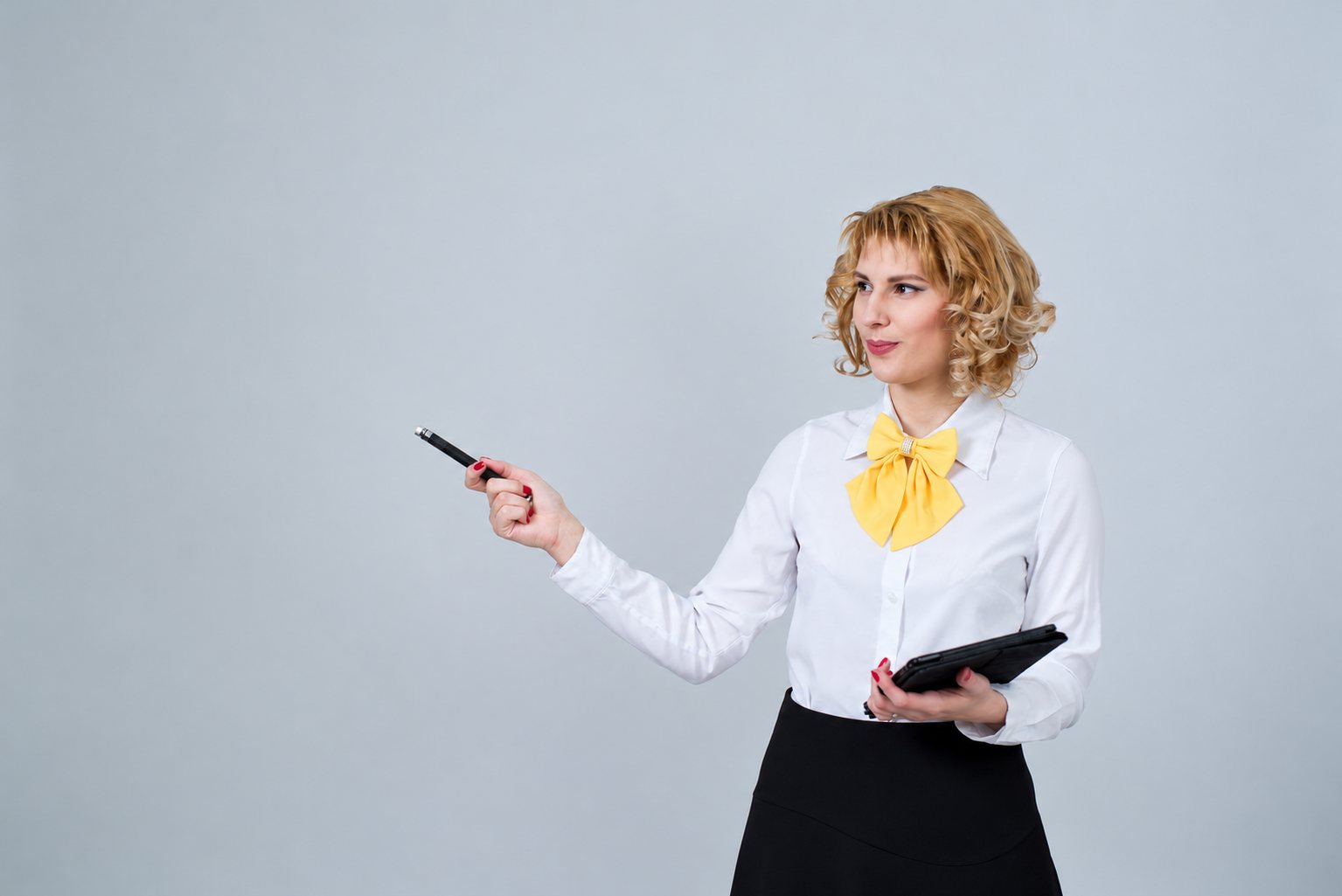 Une femme blonde aux cheveux bouclés, vêtue d'un chemisier blanc avec un nœud jaune, pointe un stylo vers la gauche en tenant une tablette.