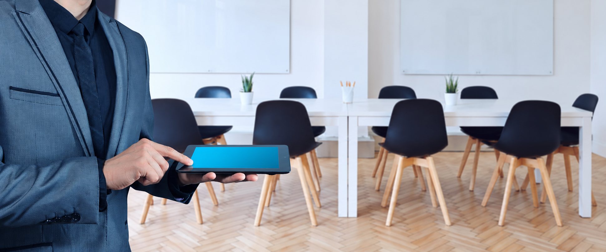 Un homme en costume bleu touche l'écran bleu d'une tablette devant une salle de réunion lumineuse avec une table blanche et des chaises sombres.