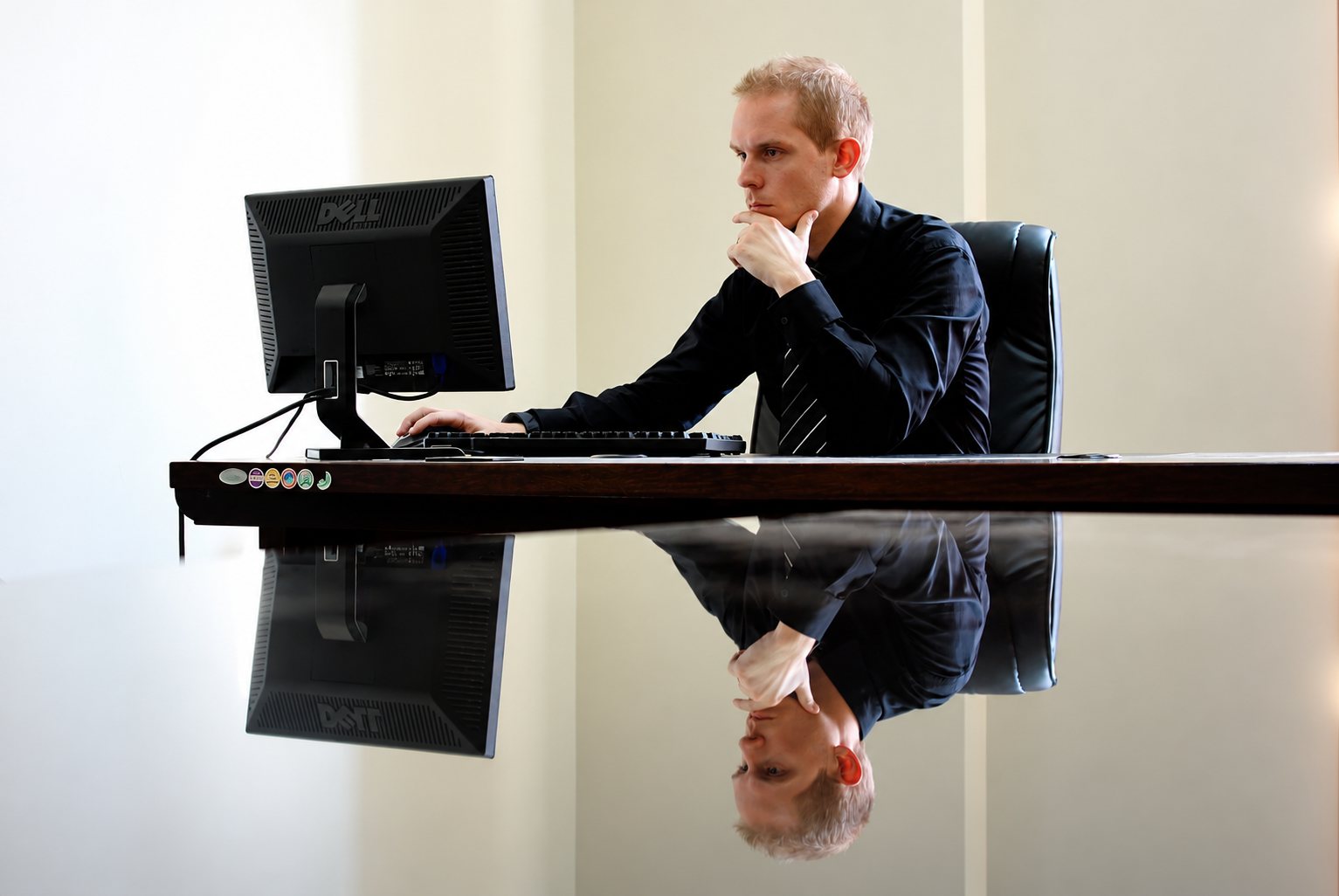 Un homme en chemise noire, la main sous le menton, regarde un écran d'ordinateur Dell sur un bureau laqué qui reflète son image.